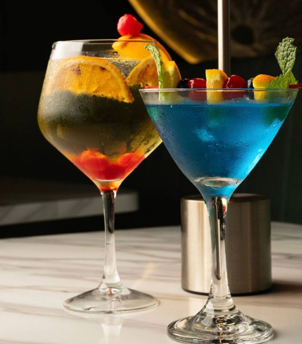 Dos cócteles en la barra de mármol: uno dorado con naranja y uno azul con frutas y menta